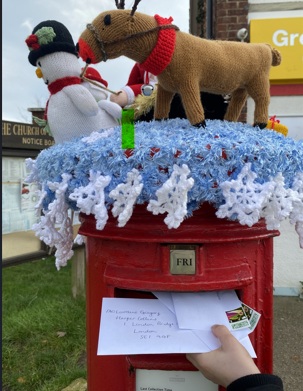Child posting letters to their favourite authors into a village post box adorned with a knitted festive topper