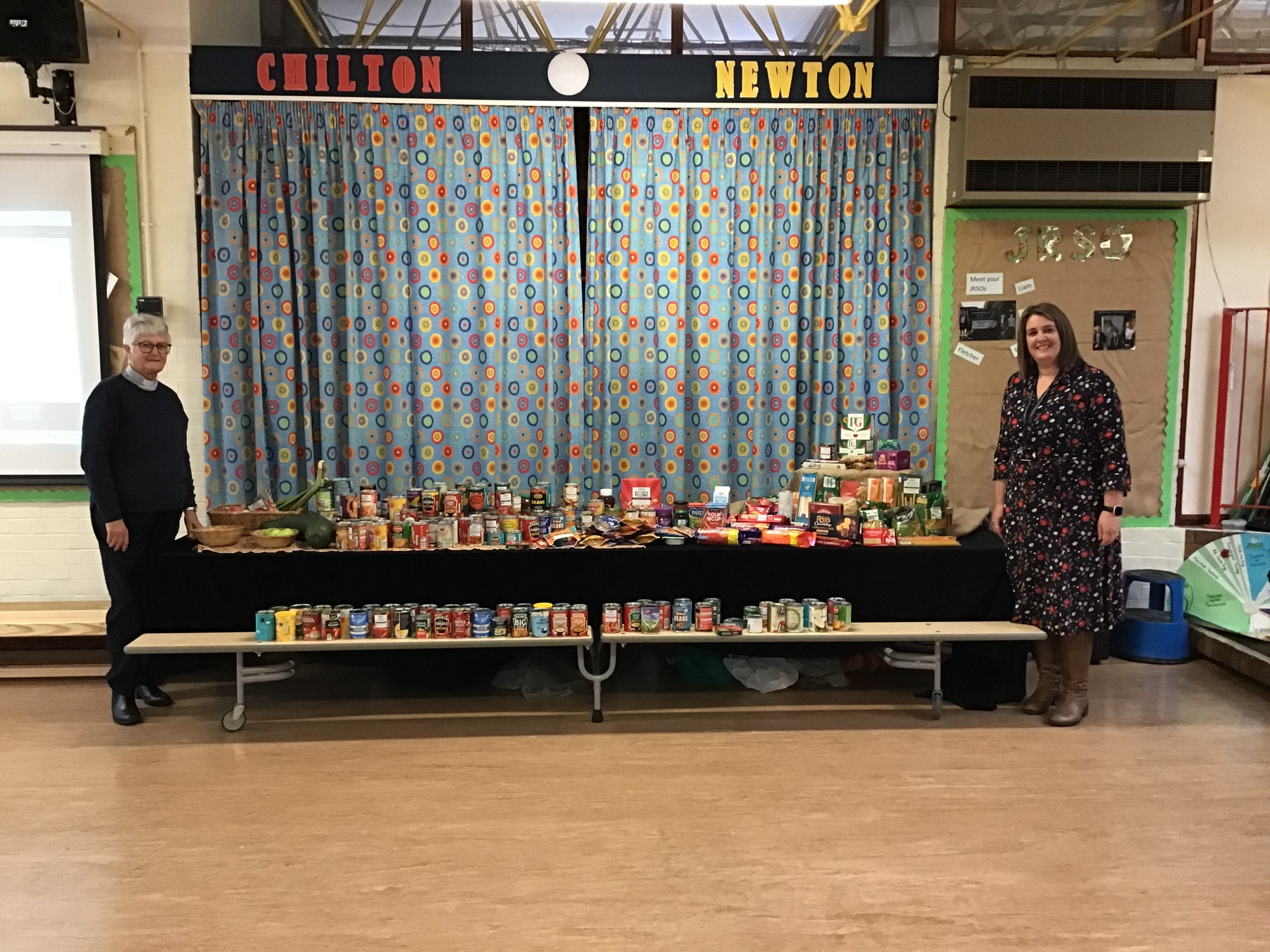 Reverend Faith and Headteacher Mrs Baker standing by a table displaying all of the wonderful Harvest Festival food donations brought in by the children